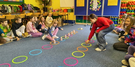 Powiększ grafikę: Dzieci siedzą na dywanie w dwóch rzędach na przeciwko siebie. Między nimi ułożone są dwa rzędy kolorowych kółek z kubkami w środku. Dwie dziewczynki rywalizują składając pojedynczo kubki.