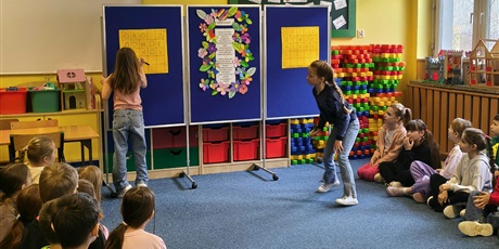 Powiększ grafikę: Dwie dziewczynki rysują znaki na pomarańczowych planszach. Plansze wiszą na granatowej tablicy. Pozostałe dzieci siedzą na dywanie w świetlicy.