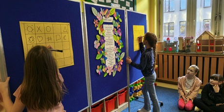 Powiększ grafikę: Dwie dziewczynki rysują znaki na pomarańczowych planszach. Plansze wiszą na granatowej tablicy.