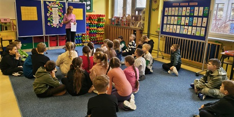 Powiększ grafikę: Dzieci siedzą na dywanie w świetlicy zwrócone w stronę nauczycielki. Nauczycielka stoi obok granatowej tablicy, na której wisi laurka z życzeniami i kolorowymi kwiatami z okazji Dnia Kobiet.