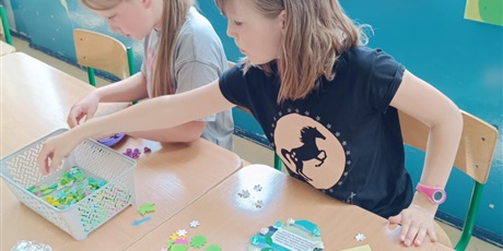 Powiększ grafikę: Dwie dziewczynki siedzą  przy stolikach i dekorują drewniane serca dla swoich mam.