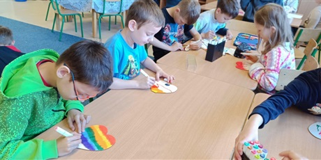 Powiększ grafikę: Grupa dzieci siedzi  przy stolikach i dekoruje drewniane serca dla swoich mam.