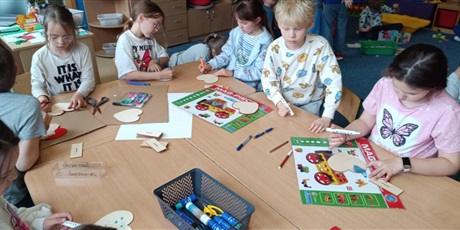 Powiększ grafikę: Grupa dzieci siedzi  przy stolikach i dekoruje drewniane serca dla swoich mam.