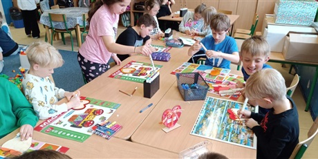 Powiększ grafikę: Grupa dzieci siedzi  przy stolikach i dekoruje drewniane serca dla swoich mam.