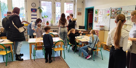 Powiększ grafikę: Przyszli uczniowie szkoły odwiedzają wraz z rodzicami szkolne zerówki, dzieci bawią się przy stolikach a rodzice rozmawiają z nauczycielkami. 