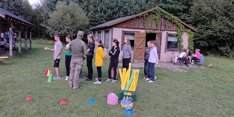 Powiększ grafikę: Na trawiastym boisku dzieci rzucają piłką do celu. Mężczyzna w wojskowym stroju udziela im instrukcji. W tle dziewczynki siedzą na ławce.