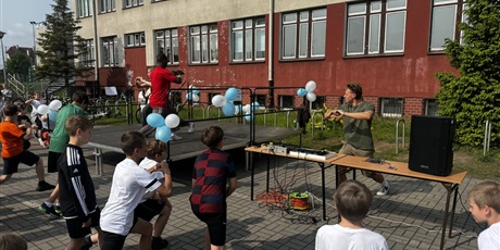 Powiększ grafikę: Grupa ubranych na sportowo dzieci w wieku szkolnym ćwiczy na boisku. W tle widać osoby dorosłe pokazujące elementy ruchu.