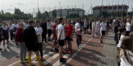 Powiększ grafikę: Grupa dzieci w starszym wieku szkolnym znajduje się na boisku szkolnym. Są ubrani na sportowo. Uczniowie są uśmiechnięci.