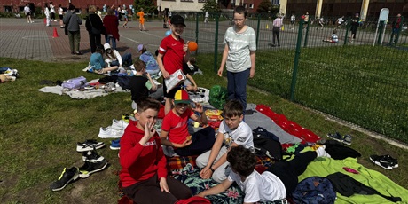 Powiększ grafikę: Grupa chłopców siedzi i stoi na kocu. Koc leży na trawie. Dzieci uśmiechają się i machają do aparatu. Mają zdjęte buty. W tle widać inne osoby dorosłe.