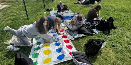 Powiększ grafikę: Dziewczynki w wieku szkolnym grają w grę Twister. Mają dłonie ułożone na kolorowych kółkach na planszy. Inne dzieci znajdują się w tle. Siedzą na kocu na trawie.