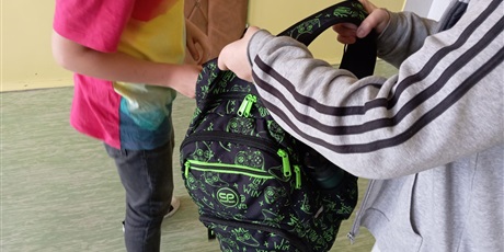 Powiększ grafikę: Uczeń podaje drugiemu uczniowi plecak.
