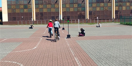 Powiększ grafikę: Dwoje dzieci jedzie rowerami przez szkolne miasteczko ruchu drogowego. Przejeżdżają obok sygnalizacji świetlnej i wyznaczonych pasów.