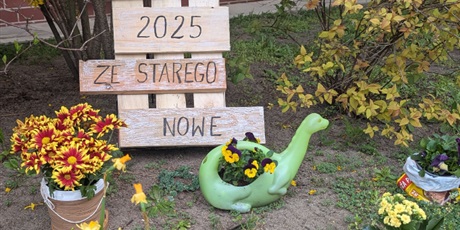 Powiększ grafikę: Napis na kawałkach desek: „ZE STAREGO NOWE, DZIEŃ ZIEMI 2025”, kolorowe kwiaty w różnych pojemnikach (kobiałce, wiaderku, konewce, torebce po kawie). Wszystko ustawione na trawie.