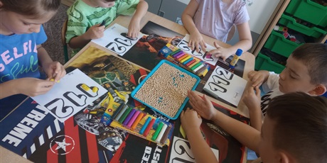Powiększ grafikę: Grupa dzieci sześcioletnich siedzi przy stoliku i przy użyciu plasteliny i kulek grochu ozdabiają nr alarmowy 112. Dzieci pracują w skupieniu.