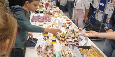 Powiększ grafikę: Potrawy z różnych krajów europejskich zaprezentowane na stolikach na holu głównym szkoły. Wokół uczniowie wybierający potrawy, aby je zakupić.