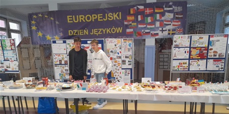 Powiększ grafikę: Dwóch uczniów przygotowuje stanowisko z potrawami, w tyle widać baner z napisem Europejski Dzień Języków Obcych oraz dekorację z plakatów o krajach europejskich.