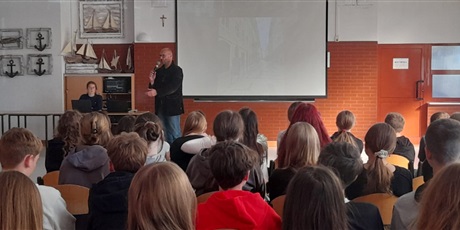 Powiększ grafikę: Nauczyciel prowadzi spotkanie - Nauczyciel stoi przed siedzącą grupą uczniów, w ręku trzyma mikrofon, w tle widać prezentację na dużym białym ekranie.