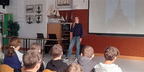 Powiększ grafikę: Wiktoria Syrytczyk - Młoda dziewczyna stoi z mikrofonem przed siedzącą grupą uczniów, w tle widać prezentację na dużym białym ekranie.