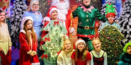 Powiększ grafikę: Grupa uczniów i nauczycieli przebranych świątecznie pozuje z Mikołajem oraz Śnieżynką  na tle choinek. Dzieci przebrane są za choinki.