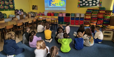 Powiększ grafikę: Grupa dzieci siedzi na dywane tyłem do obiektywu. Dzieci wpatrują się w ekran przedstawiający  film pt. "Siedem Kontynentów Świata".