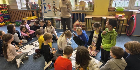 Powiększ grafikę: Dwie grupy dzieci siedzą na dywanie w świetlicy. Między nimi stoi nauczycielka trzymająca w rękach małe karteczki. Dzieci patrzą w jej kierunku.