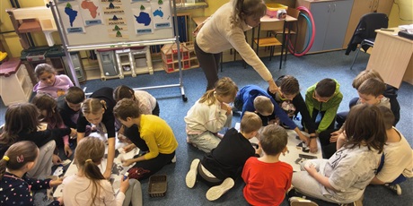 Powiększ grafikę: Dwie grupy dzieci siedzą na dywanie w świetlicy. Pochylają się nad plakatami znajdującymi się na podłodze przyklejając karteczki. Nauczycielka stoi i wskazuje ręka na plakat.