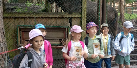 Powiększ grafikę: Grupa siedmioletnich dzieci  pozuje do zdjęcia. Dzieci są ubrane letnio, mają czapki na głowach. W tle widać ogrodzenie zoo wraz ze zwierzęciem - łosiem
