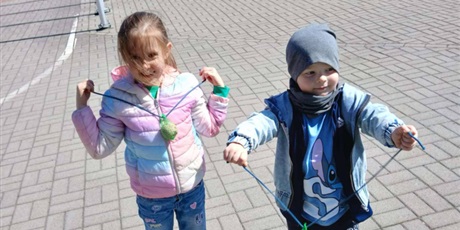 Powiększ grafikę: Na zdjęciu dwoje 6-letnich dzieci uśmiecha się. Dziewczynka ma ubraną kurtkę. Chłopiec ma nałożoną czapkę i kurtkę. Oboje pokazują pokarm dla ptaków w kształcie kuli w zielonej siatce.