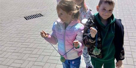 Powiększ grafikę: Na zdjęciu dwoje uśmiechniętych dzieci sześcioletnich niesie w rękach pokarm dla ptaków. Dzieci są na podwórku. Mają na sobie kurtki.