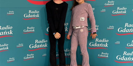 Powiększ grafikę: Dwie dziewczynki w wieku 7 lat pozują do zdjęcia na tle ścianki z napisem Radio Gdańsk. Napis jest koloru białego, pod nim znajduje się czerwona fala. Wszystko jest na turkusowym tle. Dziewczynki uśmi