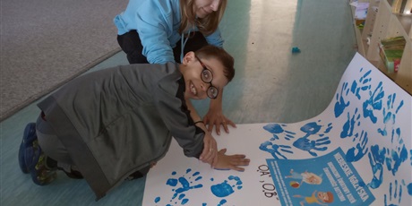 Powiększ grafikę: chłopiec w okularach z pomocą pani nauczycielki odbija swoją dłoń na plakacie. Pani nauczycielka ma na sobie niebieską bluzę. Chłopiec patrzy w kadr, uśmiecha się. Jego buza jest koloru szarego.