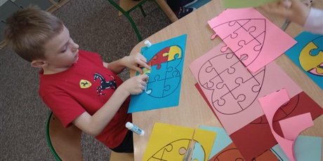 Powiększ grafikę: Chłopiec w czerwonej koszulce dopasowuje nakleja zielony puzzel  na niebieskiej kartce. Na stole leżą różnokolorowe kartki.