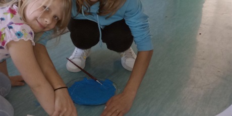 Powiększ grafikę: Uśmiechnięta dziewczynka w blond włosach odbija swoją dłoń na grupowym plakacie.