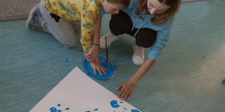 Powiększ grafikę: Chłopiec w żółtej bluzie w kolorowe auta wkłada dłoń w niebieską farbę. Na podłodze widać plakat z odbitymi niebieskimi dłońmi dzieci.
