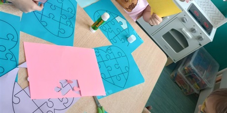 Powiększ grafikę: Dzieci w wieku lat 7 pracują przy stole. Dziewczynka wycina potrzebny element. Chłopiec dopasowuje puzzel. Widać różnokolorowych kartki.