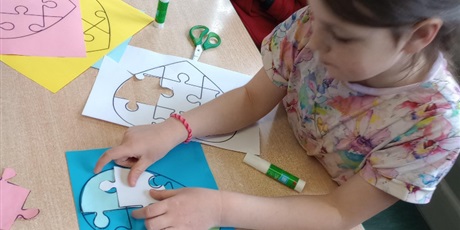 Powiększ grafikę: Chłopiec i dziewczynka pracują w skupieniu. Dziewczynka w ciemnych włosach dopasowuje biały puzzel do szablonu. Chłopiec w pomarańczowej bluzie trzyma w dłoni czerwoną kartkę i wycina.