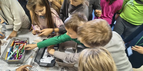 Powiększ grafikę: Grupa dzieci stoi dookoła stołu. Dzieci biorą karteczki i mazaki z pojemników znajdujących się na stole.