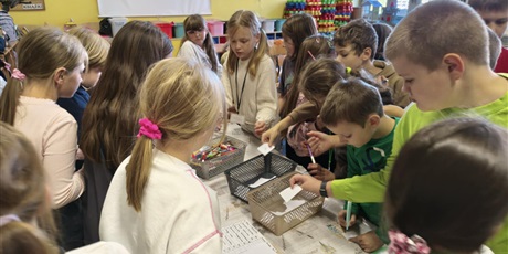 Powiększ grafikę: Grupa dzieci stoi dookoła stołu. Dzieci biorą karteczki i mazaki z pojemników znajdujących się na stole.