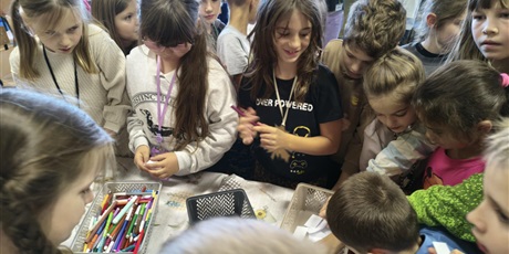 Powiększ grafikę: Grupa dzieci stoi dookoła stołu. Dzieci biorą karteczki i mazaki z pojemników znajdujących się na stole.