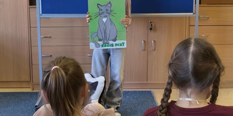 Powiększ grafikę: Dziewczynka  stoi zwrócona przodem do publiczności.   W ręku trzyma obraz kota.  Za nią stoi granatowa tablica z kolorową dekoracją i napisem I Edycja Konkursu Recytatorskiego „W kręgu poezji Ludwika 