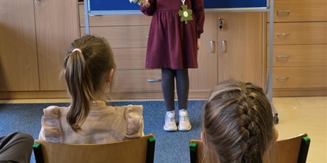 Powiększ grafikę: Dziewczynka w bordowej sukience stoi zwrócona przodem do publiczności. W ręku trzyma małą zieloną maskotkę.  Za nią stoi granatowa tablica z kolorową dekoracją i napisem I Edycja Konkursu Recytatorski