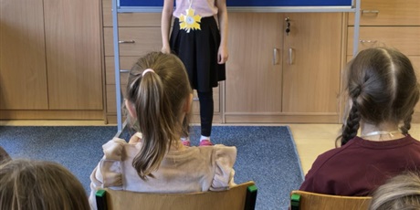 Powiększ grafikę: Dziewczynka w różowej  bluzce i czarnej spódnicy stoi zwrócona przodem do publiczności.  Za nią stoi granatowa tablica z kolorową dekoracją i napisem I Edycja Konkursu Recytatorskiego.