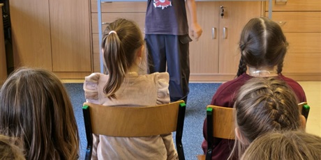 Powiększ grafikę: Dziewczynka w ciemnej bluzce z aplikacją kota stoi zwrócona przodem do publiczności.  Za nią stoi granatowa tablica z kolorową dekoracją i napisem I Edycja Konkursu Recytatorskiego „W kręgu poezji Lud