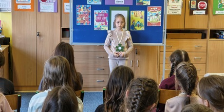 Powiększ grafikę:  Dziewczynka w jasnej bluzce i okularach stoi zwrócona przodem do publiczności.  Za nią stoi granatowa tablica z kolorową dekoracją i napisem I Edycja Konkursu Recytatorskiego „W kręgu poezji Ludwika 