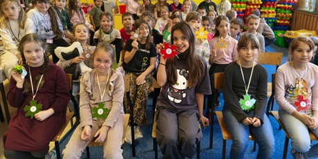 Powiększ grafikę: Grupa dzieci pozuje do zdjęcia siedząc na krzesłach i ławkach. Dziewczynki w dwóch pierwszych rzędach mają na szyi zawieszone numerki.