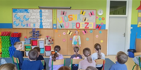 Powiększ grafikę: Grupa dzieci siedzi tyłem.  Dziewczynka stoi przodem i recytuje wiersz.