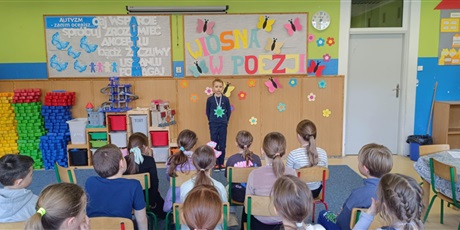 Powiększ grafikę: Grupa dzieci siedzi tyłem.  Chłopiec stoi przodem i recytuje wiersz