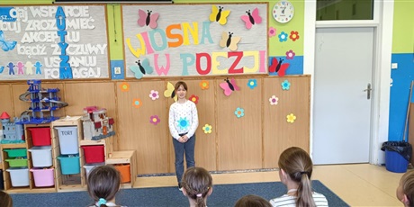 Powiększ grafikę: Grupa dzieci siedzi tyłem.  Dziewczynka stoi przodem i recytuje wiersz