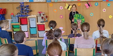 Powiększ grafikę: Grupa dzieci siedzi tyłem.  Dziewczynka stoi przodem i recytuje wiersz. Dziewczynka trzyma w ręku drzewo z papieru.