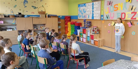 Powiększ grafikę: Grupa dzieci siedzi na krzesełkach.  Dziewczynka stoi przed dziećmi i recytuje wiersz.